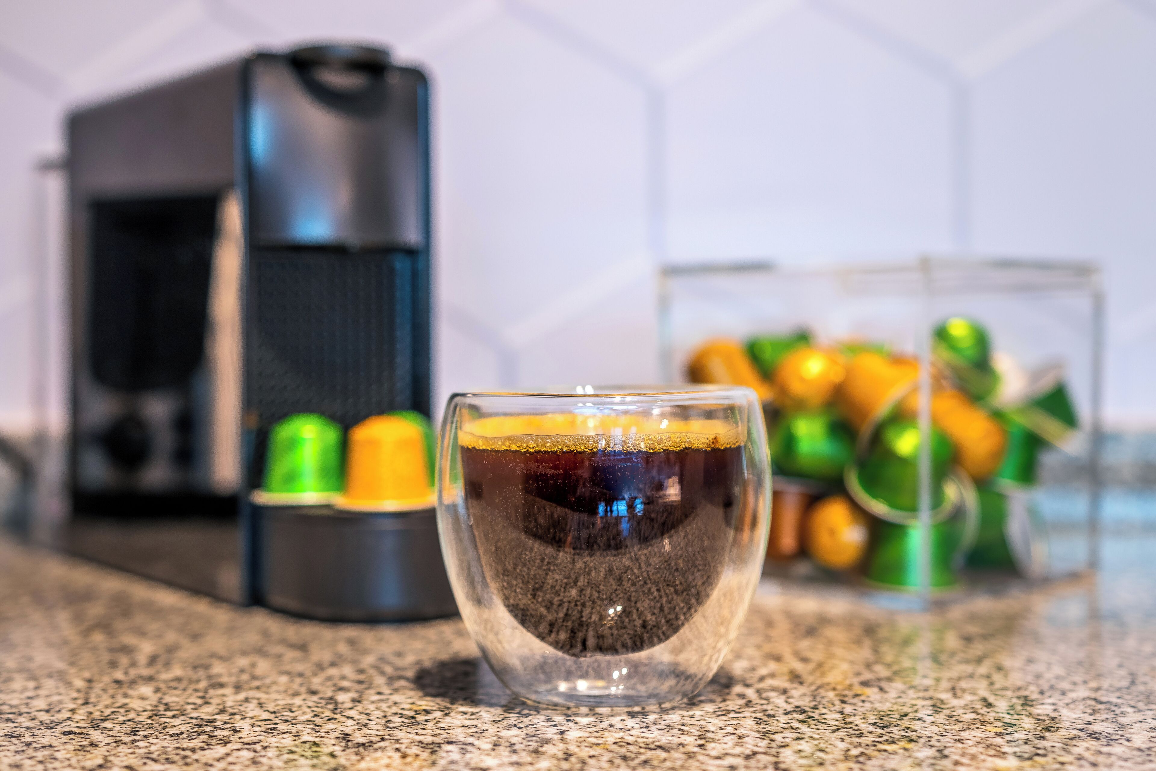 Una taza de café sobre una encimera, con una cafetera y un recipiente de cápsulas de café al fondo.