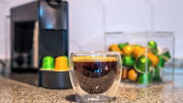 Una taza de café sobre una encimera, con una cafetera y un recipiente de cápsulas de café al fondo.