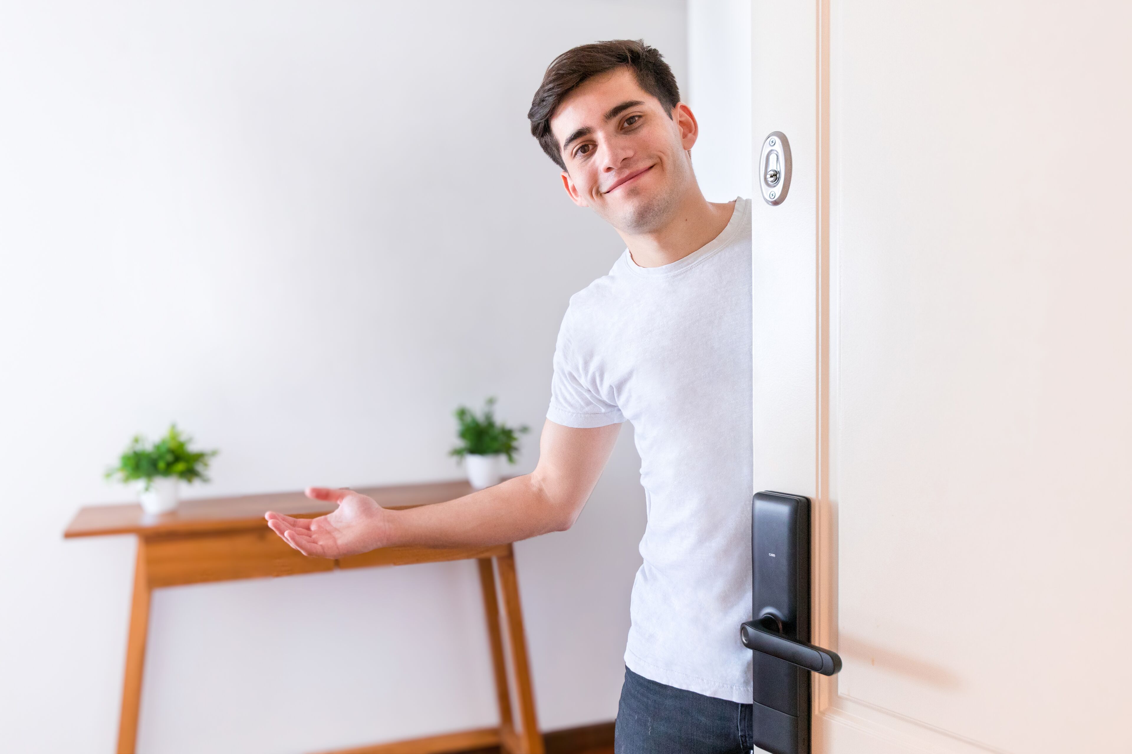 A person standing at a door with a modern handle, a wooden desk, and two potted plants in the background.