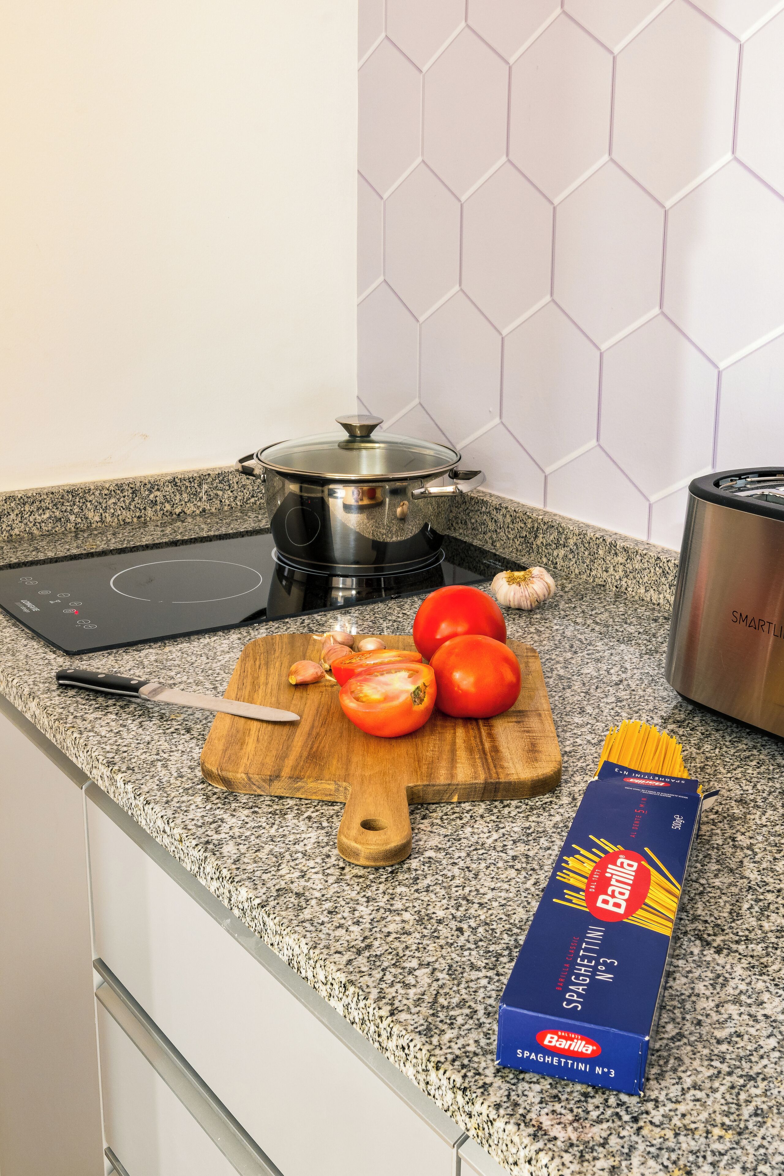 A kitchen counter with a box of Barilla spaghetti, a cutting board with tomatoes and garlic, a pot on an induction cooktop, and a toaster.