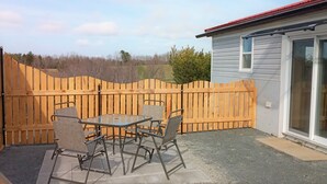 A patio area with a table and chairs, a wooden fence, and a house with a red roof and sliding glass doors. - Red Barn Bed & Breakfast (Lunenburg)