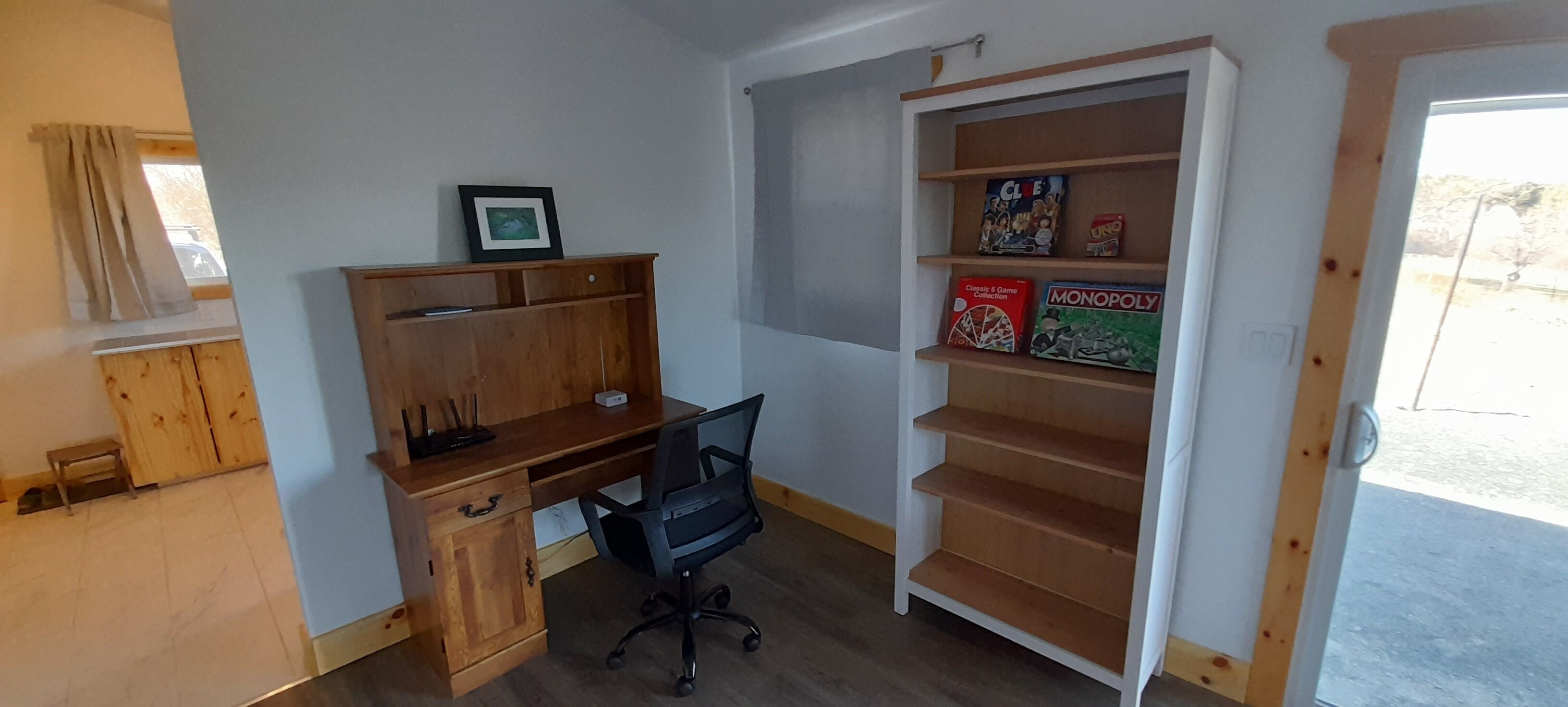 Una habitación con un escritorio de madera, una silla de oficina negra, una estantería con juegos de mesa y una ventana con cortinas.