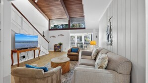 Un salón amplio con un televisor grande, asientos cómodos, techo de madera y vistas al exterior.