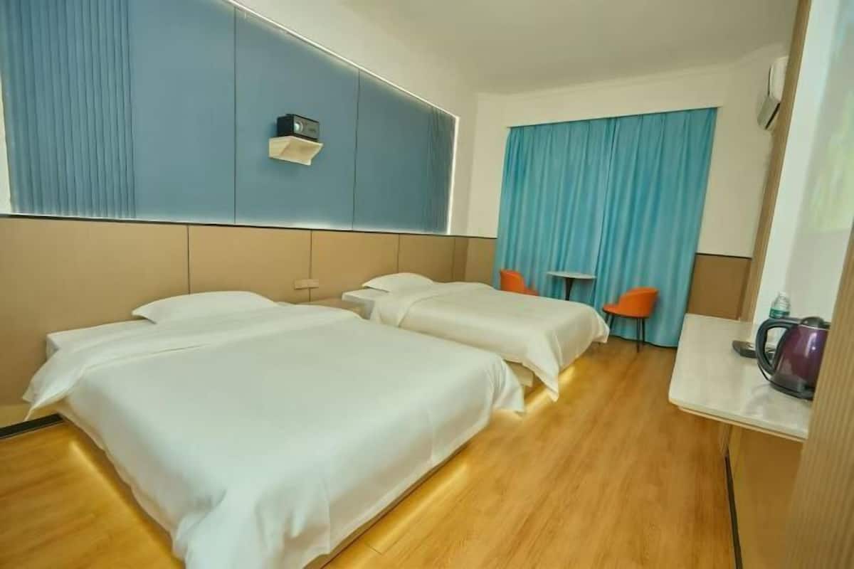 A hotel room with two beds, a desk, a chair, a window with curtains, and a wall-mounted radio.