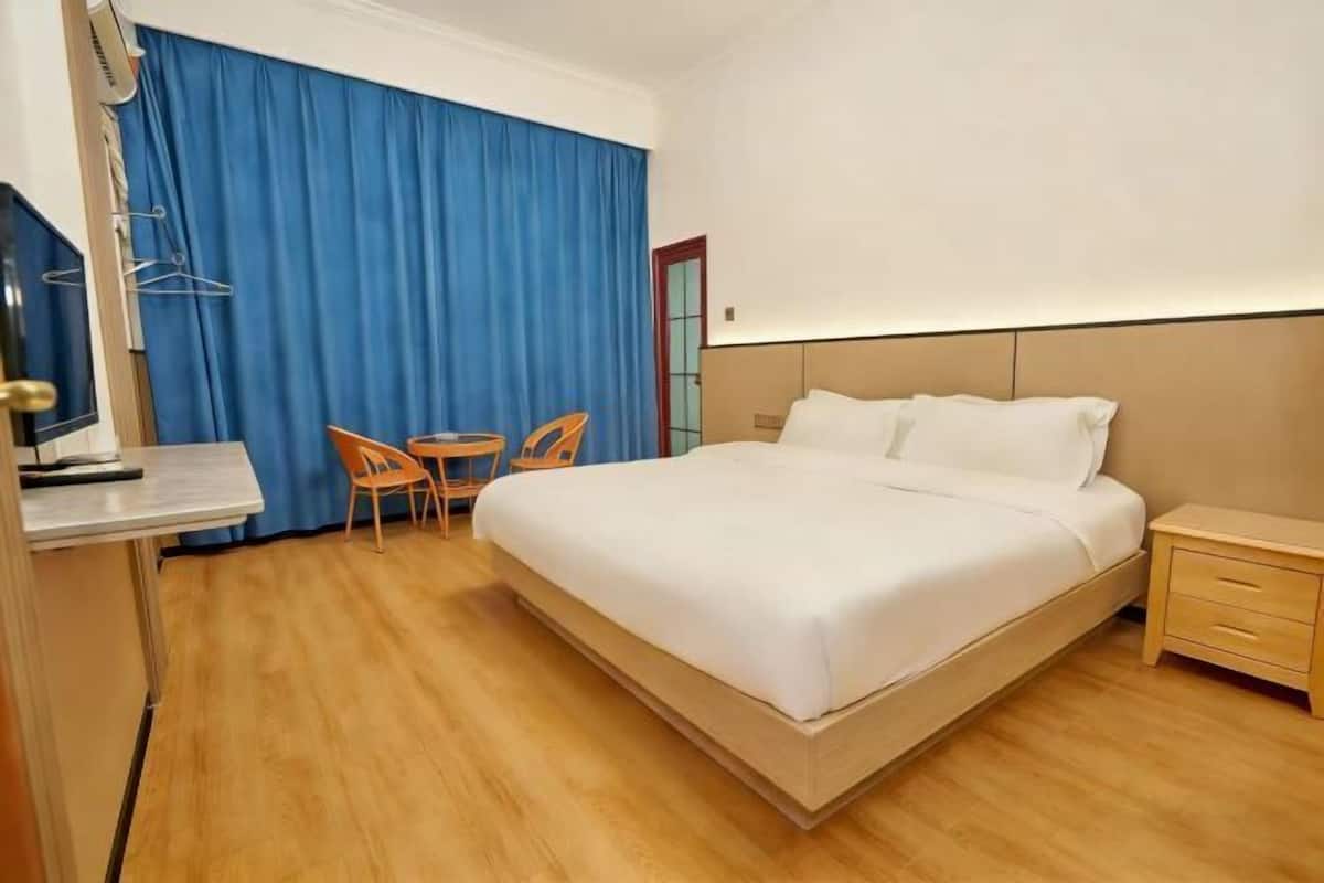 A hotel room with a large bed, a desk with a chair, a small table, and a blue curtain.
