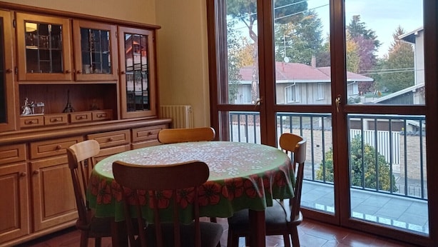 Een eethoek met een ronde tafel, afgedekt met een tafelkleed met bloemenmotief, houten stoelen en een kast met glazen deuren.