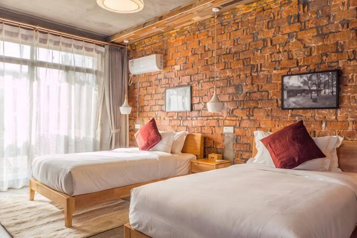 Een kamer met twee bedden, een bakstenen muur, een raam met gordijnen en een schilderij aan de muur.