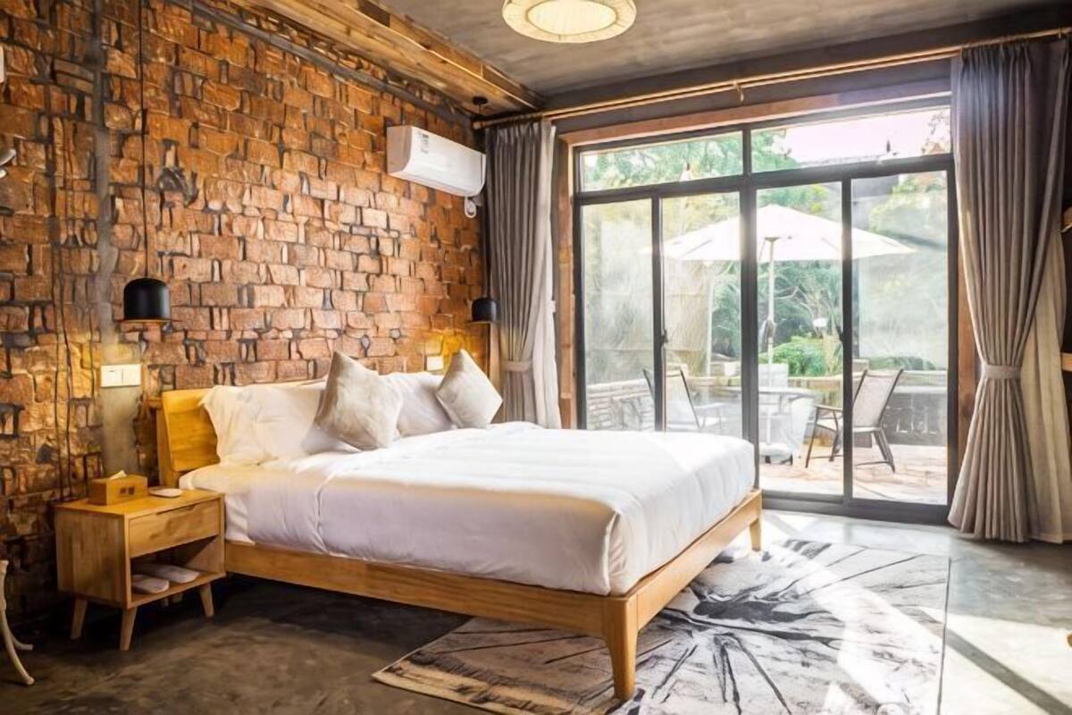 Een slaapkamer met een bakstenen muur, een groot bed, een nachtkastje, een lamp en uitzicht op een terras.