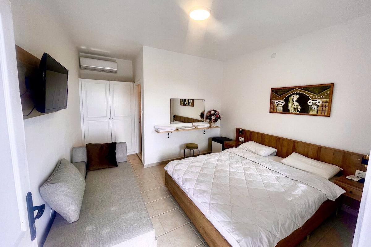 A hotel room with a bed, a bench, a TV, a wardrobe, a mirror, and a painting.