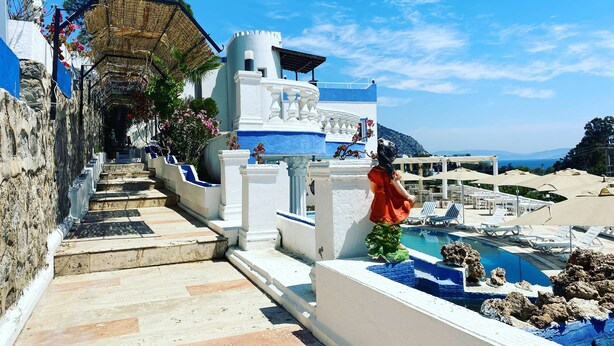 Un área junto a la piscina con una escalera de piedra que conduce a un edificio blanco con detalles azules, una estatua y vistas al mar.
