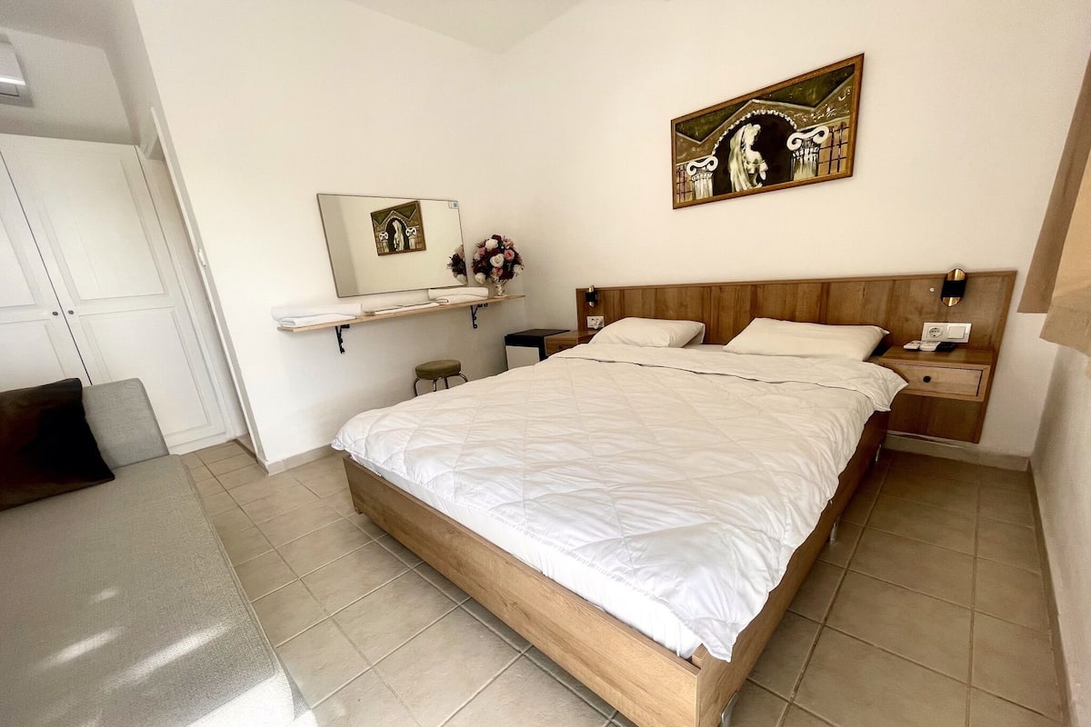 A neatly arranged bedroom with a large bed, a nightstand, a small table, a mirror, and a wall-mounted shelf with decorative items.