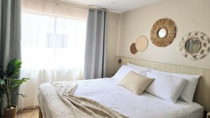 Ein ordentlich bezogenes Bett mit weißen Leinentüchern und einer beige farbenen Tagesdecke, ein Blumentopf und dekorative gewebte Wandkunst.
