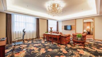 A hotel room with a large floral carpet, a chandelier, a TV mounted on the wall, and a desk with a chair.