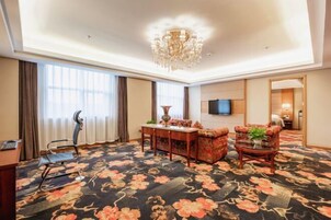Habitación de hotel con alfombra floral grande, un candelabro, televisor montado en la pared y escritorio con silla.