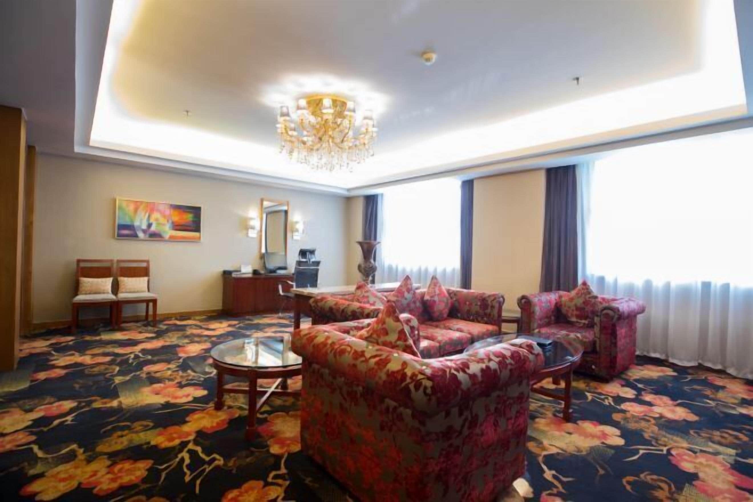 A spacious living room with floral carpet, a chandelier, and a large window with curtains.
