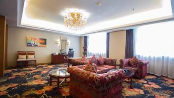 A spacious living room with floral carpet, a chandelier, and a large window with curtains.