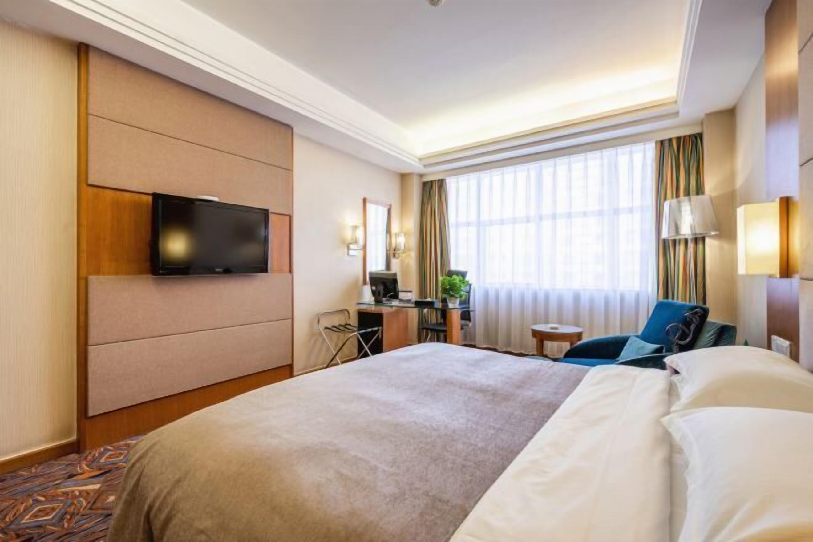 A hotel room with a large bed, a desk with a computer, a TV mounted on the wall, and a window with curtains.