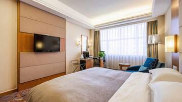 A hotel room with a large bed, a desk with a computer, a TV mounted on the wall, and a window with curtains.