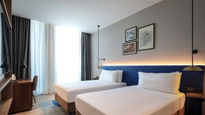 Ein Hotelzimmer mit zwei Betten, einem Schreibtisch und einem Stuhl. Zwei gerahmte Bilder hängen an der Wand.
