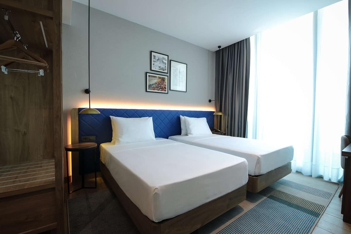 Ein Hotelzimmer mit zwei Betten, einem Holzschrank, einem Nachttisch und einem Fenster mit Vorhängen.