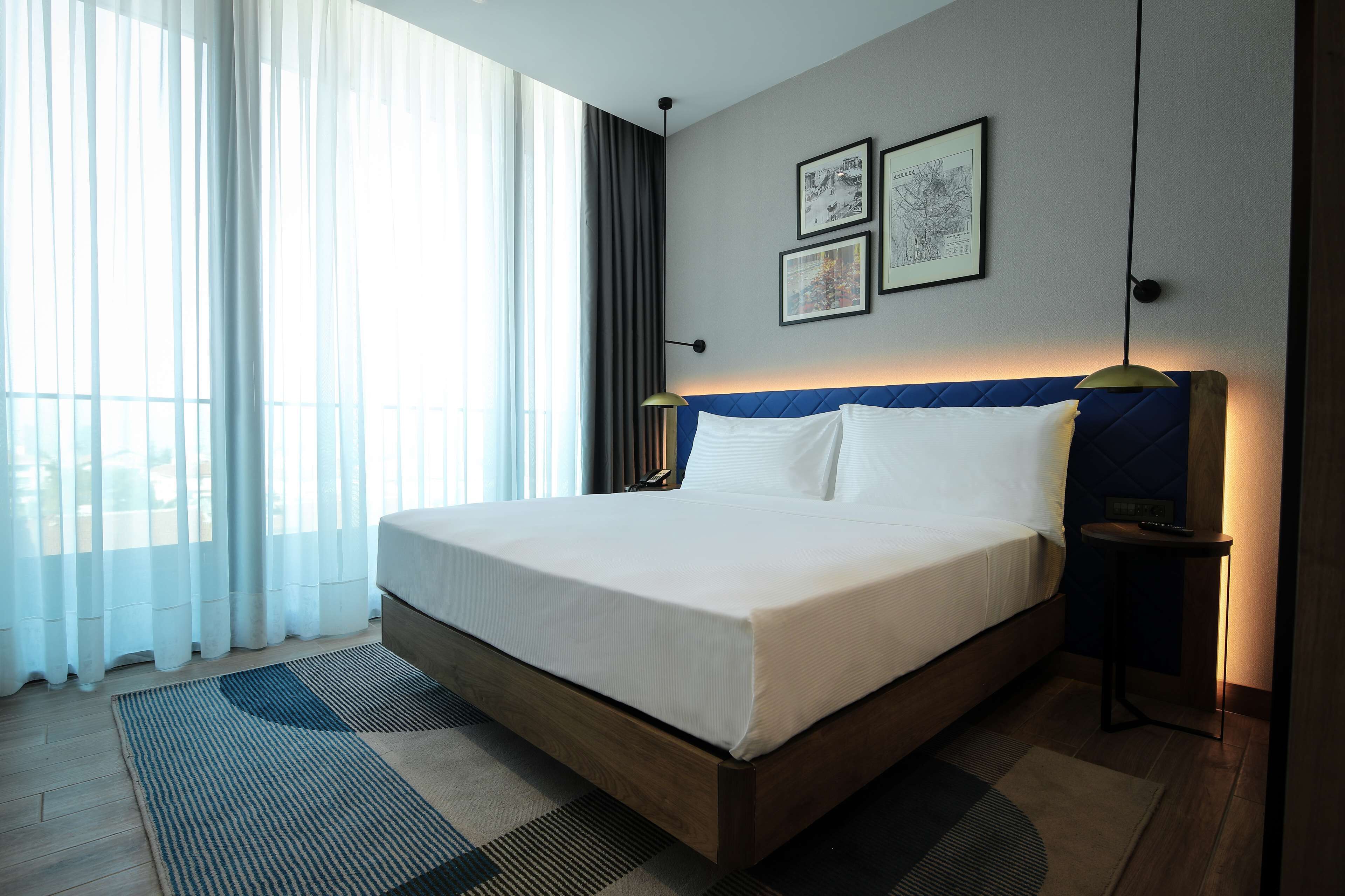 Quarto de hotel com uma cama grande, mesa de cabeceira com abajur e vista para o exterior através de cortinas finas.