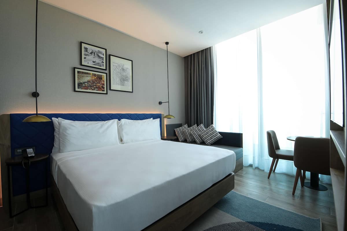 Ein modernes Hotelzimmer mit einem großen Bett, einem Schreibtisch und einem Sessel. An der Wand befinden sich gerahmte Bilder und ein Fenster mit Vorhängen.