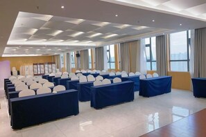 Sala de conferência com fileiras de mesas e cadeiras, janelas amplas e teto alto.