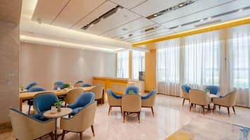 A modern dining area with a long table and multiple chairs, a reception desk, and large windows with curtains.