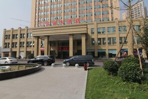 Um edifício com vários andares, entrada central, estacionamento e área ajardinada com um espelho d'água circular.