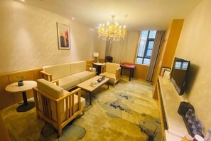 Habitación de hotel con sofá, sillones, una mesita, candelabro, televisor y ventana.