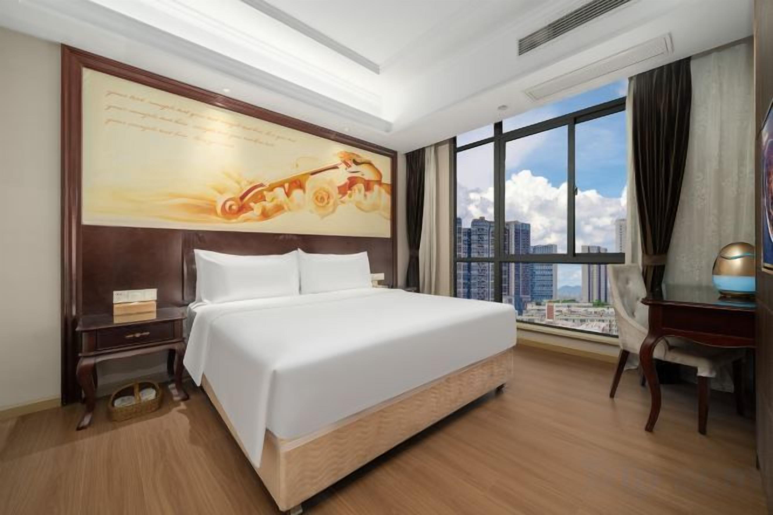 A hotel room with a large bed, a desk, a chair, a nightstand, and a large window with a city view.