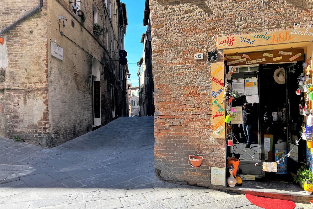 Una calle estrecha con una tienda a la derecha. La tienda tiene un letrero que enumera varios artículos, incluyendo 'caffe', 'vino' y 'aceto'. El número '12' está sobre la entrada.