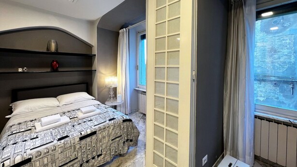 A hotel room with a bed, a nightstand, a window with curtains, and a view of the outside. - The little house on franciosa street (Siena)