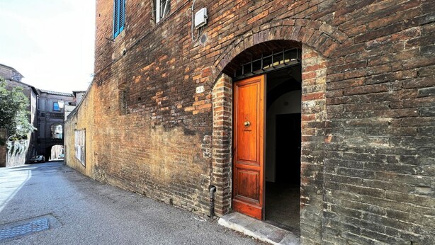 A narrow street with a brick building featuring a wooden door and an arched entrance. - Sperandie-inside the walls (Siena)