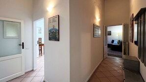 A hallway with white walls, tiled flooring, and a door on the left. There are framed pictures on the walls and a small table with chairs in the background. - Sperandie-inside the walls (Siena)