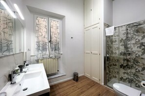 A bathroom with two sinks, a toilet, a shower with a glass door, and a trash can. - Sperandie—within the city walls (Siena)