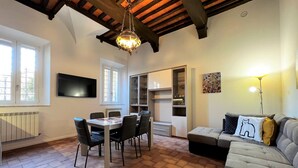A dining area with a table and chairs, a sofa, a TV mounted on the wall, and a chandelier. - Sperandie-inside the walls (Siena)