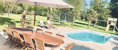 A wooden table and chairs under a large umbrella, a swimming pool, and a hammock.