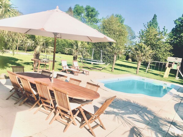 A wooden table and chairs under a large umbrella, a swimming pool, and a hammock.