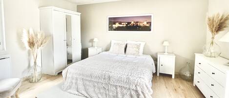 A bedroom with a large bed, white wardrobe, bedside tables with lamps, and a cityscape painting on the wall.