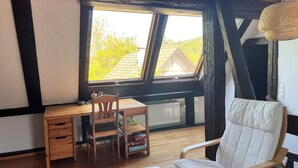 A room with a wooden desk, a chair, a cushioned seat, a window with a view of trees, and a ceiling with exposed beams. - Family-friendly apartment in an old farmhouse (Steißlingen)
