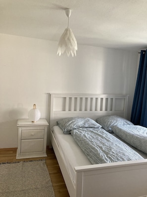 Ein Schlafzimmer mit einem weißen Bett, einem Nachttisch, einer Hängelampe und einem blauen Vorhang.
