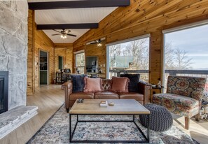 A cozy log cabin interior with a fireplace, a leather sofa, a patterned armchair, and a dining area visible through a large window.