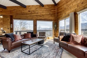 A cozy log cabin interior with leather sofas, a coffee table, and large windows offering a mountain view.