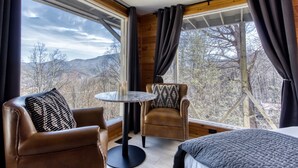 Una habitació amb un llit, una cadira de pell marró, una taula rodona amb espelmes i vistes a les muntanyes a través de grans finestres.