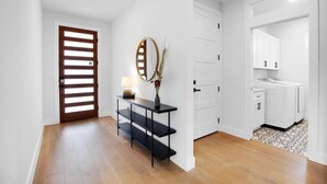 A hallway with a wooden door, a black console table with a mirror and vase, and a white door leading to a bathroom.