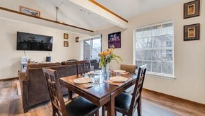 Un coin repas avec une table en bois dressée pour quatre personnes, un canapé en cuir marron et un téléviseur à écran plat fixé au mur.
