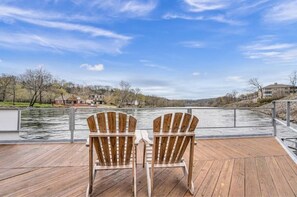 Een houten terras met twee Adirondack-stoelen gericht op een rivier, met gebouwen op de achtergrond.