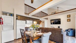 Une cuisine moderne avec des appareils électroménagers blancs, un coin repas avec une table et des chaises en bois, et un salon avec un canapé marron et un téléviseur fixé au mur.