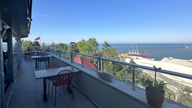 A balcony with tables and chairs, a potted plant, and a view of the sea and buildings. - Ganita Life Hotel (Trabzon)